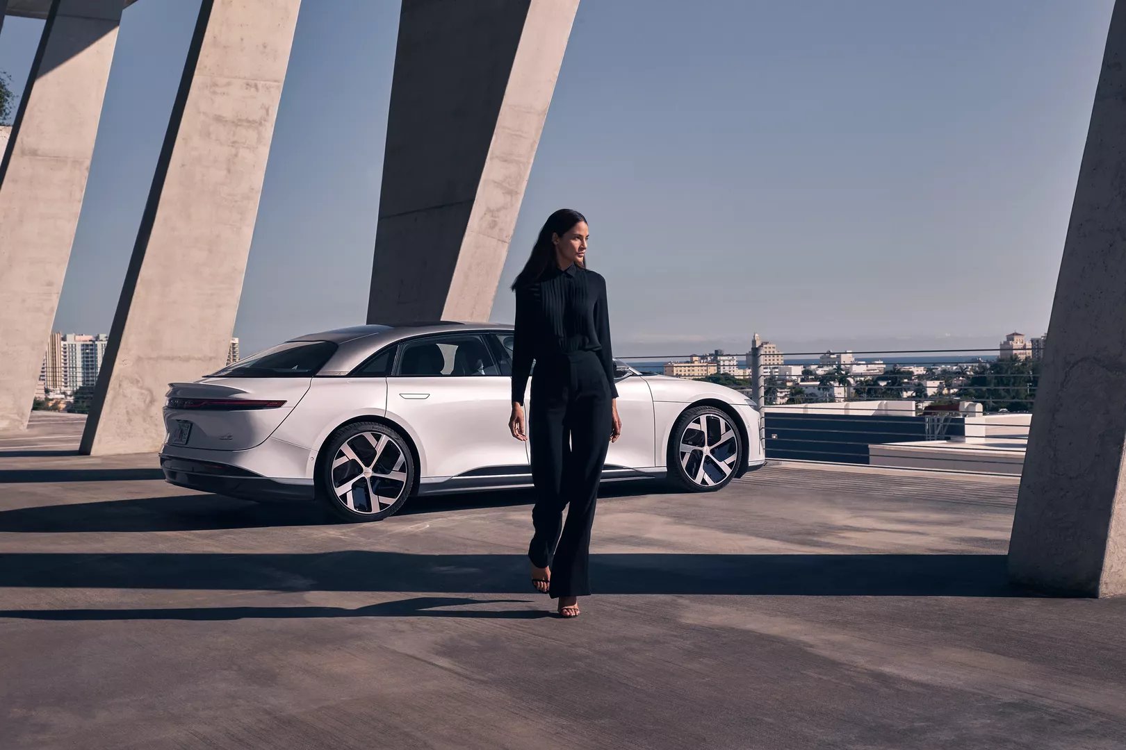 En kvinne går mot kameraet foran en Stellar White Lucid Air Dream Edition. Bilen er parkert under en moderne betongstruktur. I bakgrunnen er det bygninger og et hint av havet.