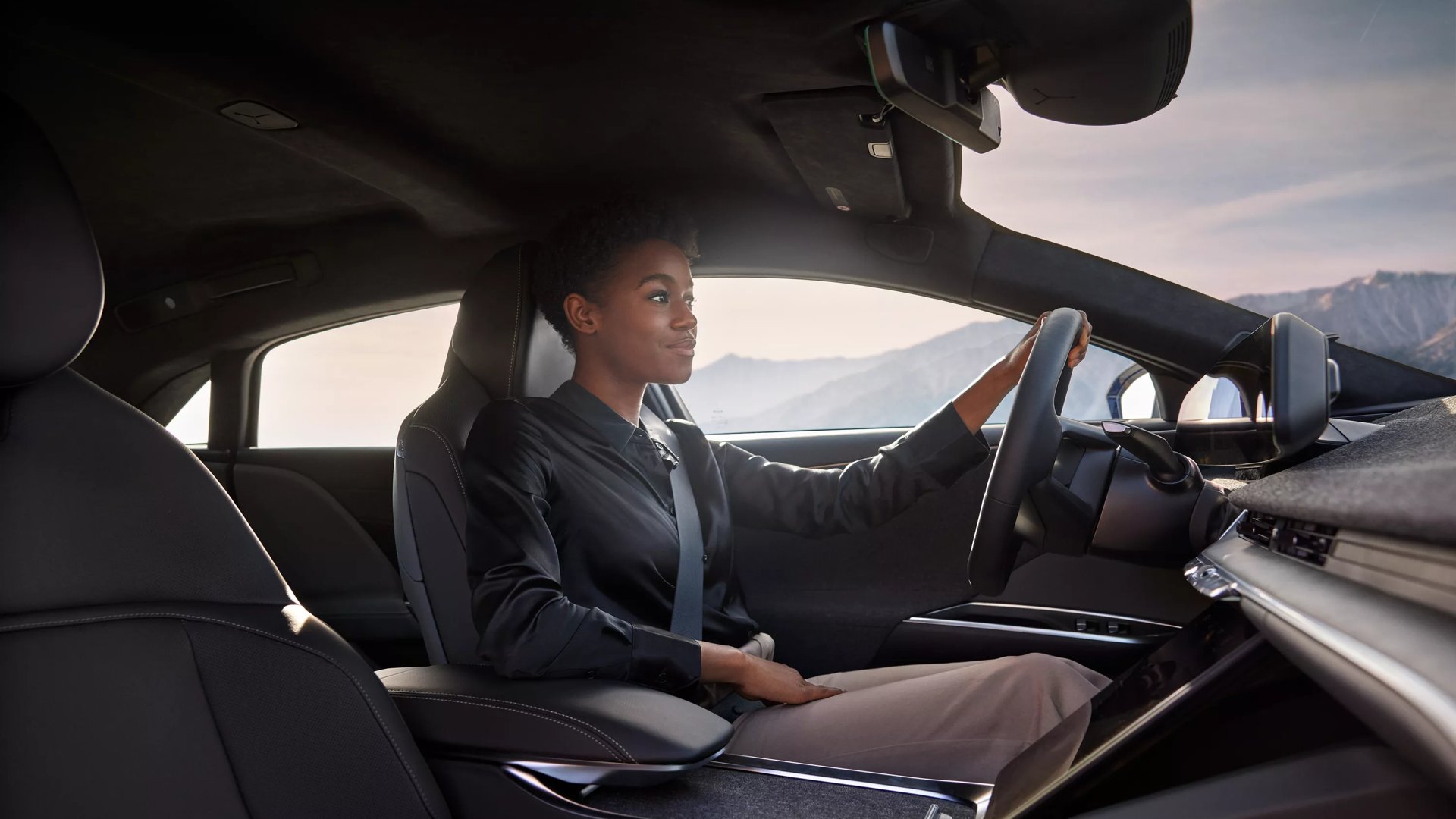 A woman is driving a Lucid Air with an air of confidence and happiness. The photo is taken from the foot well area of the passenger seat. Moutains are visible through the car windows behind the woman.
